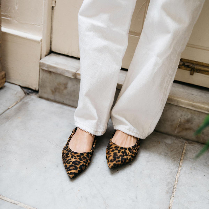 A person wears Margaux's The Roma - Leopard Suede pointed-toe ballet flats with white pants, standing on a gray tiled floor near a beige door.