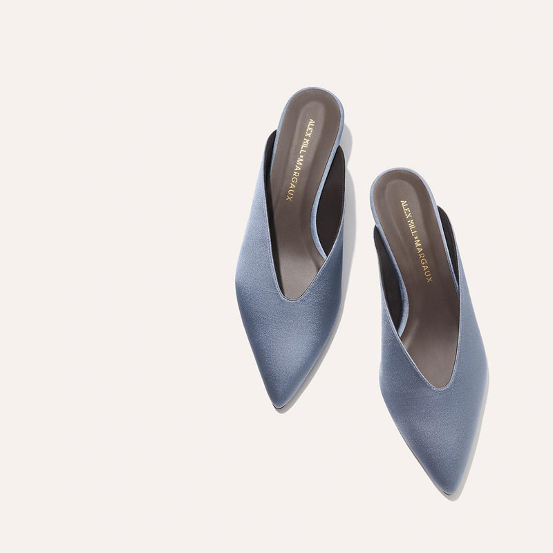 The Romy - Slate Blue Satin by Margaux are pointed-toe kitten mule slip-ons with low heels and gold-branded insoles, elegantly displayed on a light background—perfect for any occasion.