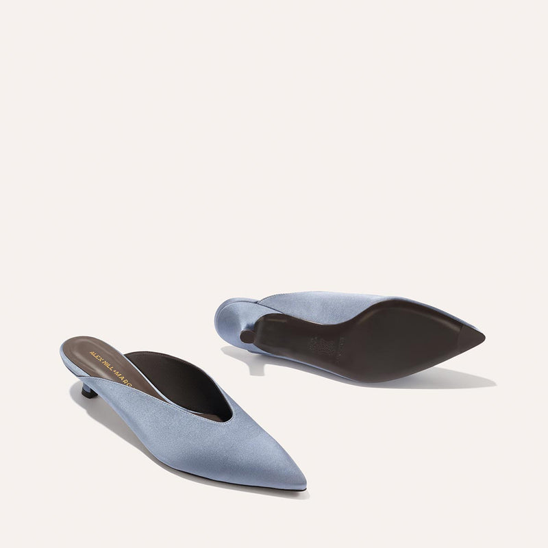 The Romy - Slate Blue Satin by Margaux: Elegant light blue pointed-toe kitten mule heels with low, slim heels. One shoe stands upright revealing the upper and insole, while the other flips to show a smooth black sole—perfect comfort for any occasion.