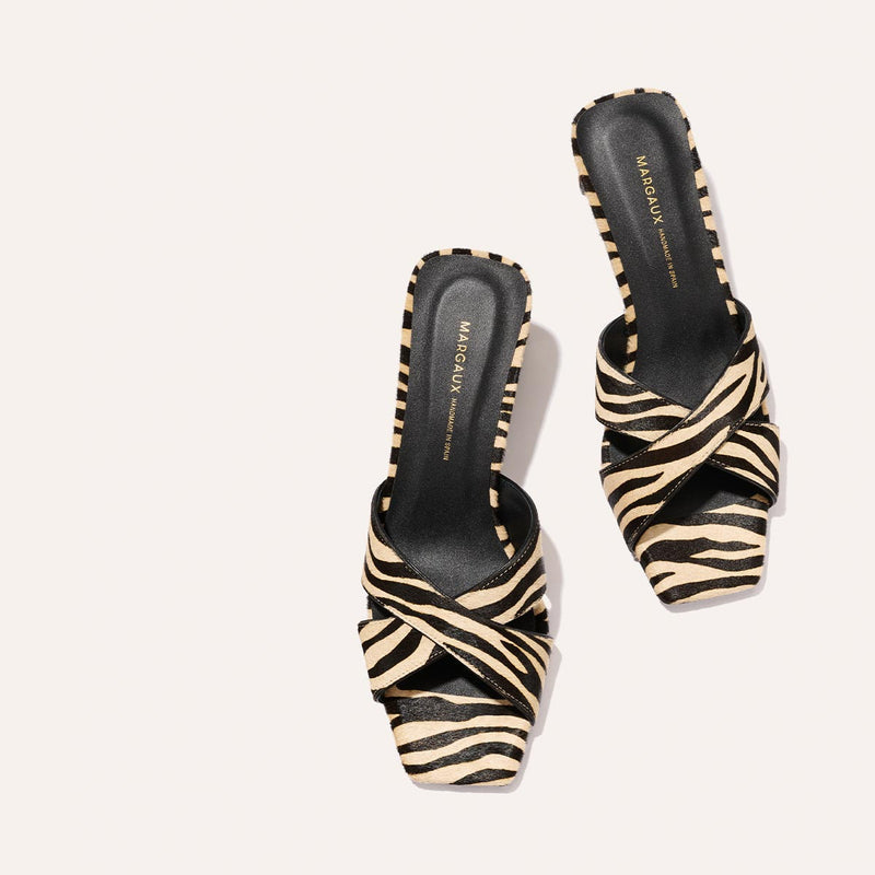 A pair of Margaux The Selene 50 - Tan Zebra Haircalf women's open-toe slip-on shoes with black and beige zebra print, crisscross straps, and black insoles displayed on a light background. The Margaux logo appears on the insoles.