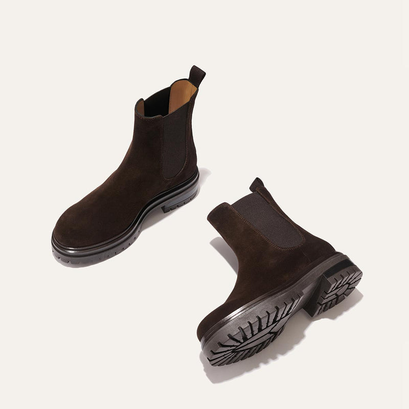 The West Chelsea Boot - Cocoa Suede by margaux-development features dark brown, water-resistant suede with black elastic panels and thick lug soles. Shown on a light background, one boot stands upright while the other lies on its side.