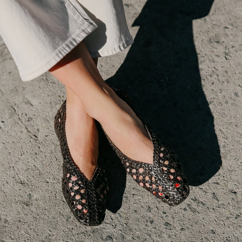 A person in high-rise cream pants wears Margaux’s The Woven Paz SS25 flats in Espresso Leather, standing on sunlit textured concrete; their toenails are painted red.