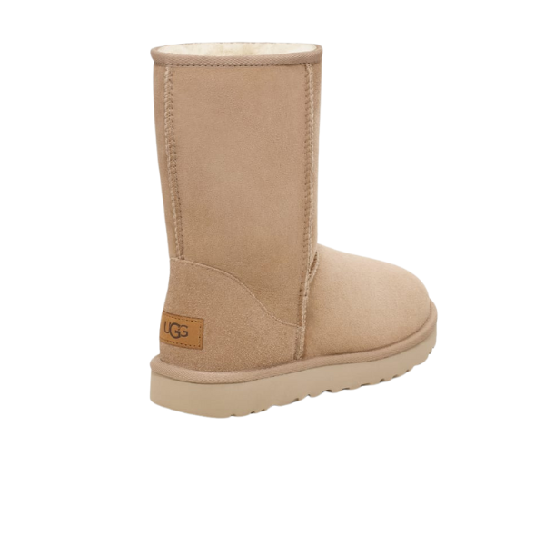 A single UGG Womens Classic Short II Boot in Sand, made of soft suede and lined with plush sheepskin for comfort, is shown mid-calf height from a rear-side angle with the UGG logo on the heel and a light-colored sole.