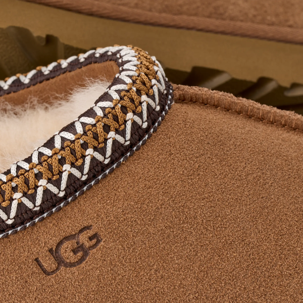 Close-up of the UGG Women's Tasman II Chestnut slipper featuring cream and tan stitching, a fuzzy white lining, and the embossed UGG logo on the side—highlighting this cozy sheepskin slipper for women.