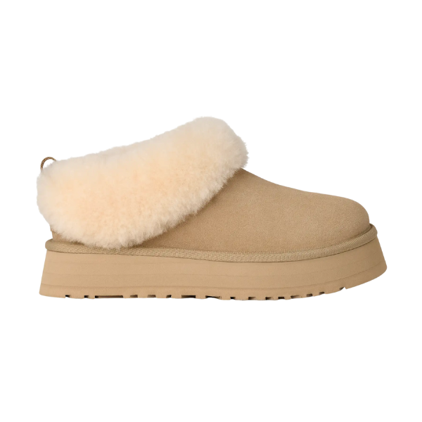 The UGG Women's Tazzelle Mustard Seed slipper features tan suede, a thick rubber sole, cream plush shearling collar, and soft sheepskin lining. Shown side view on white background. Brand: UGG.